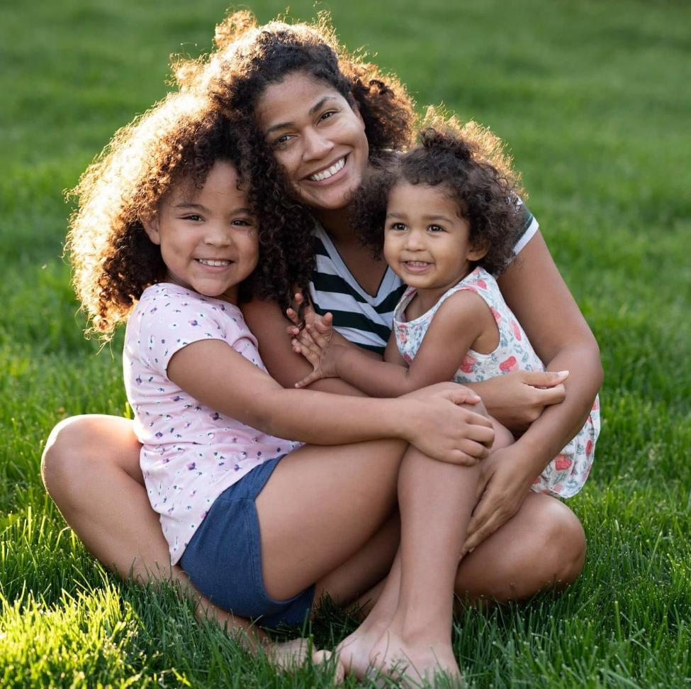 Joanna Moore sits with her two children in her lap on the grass.